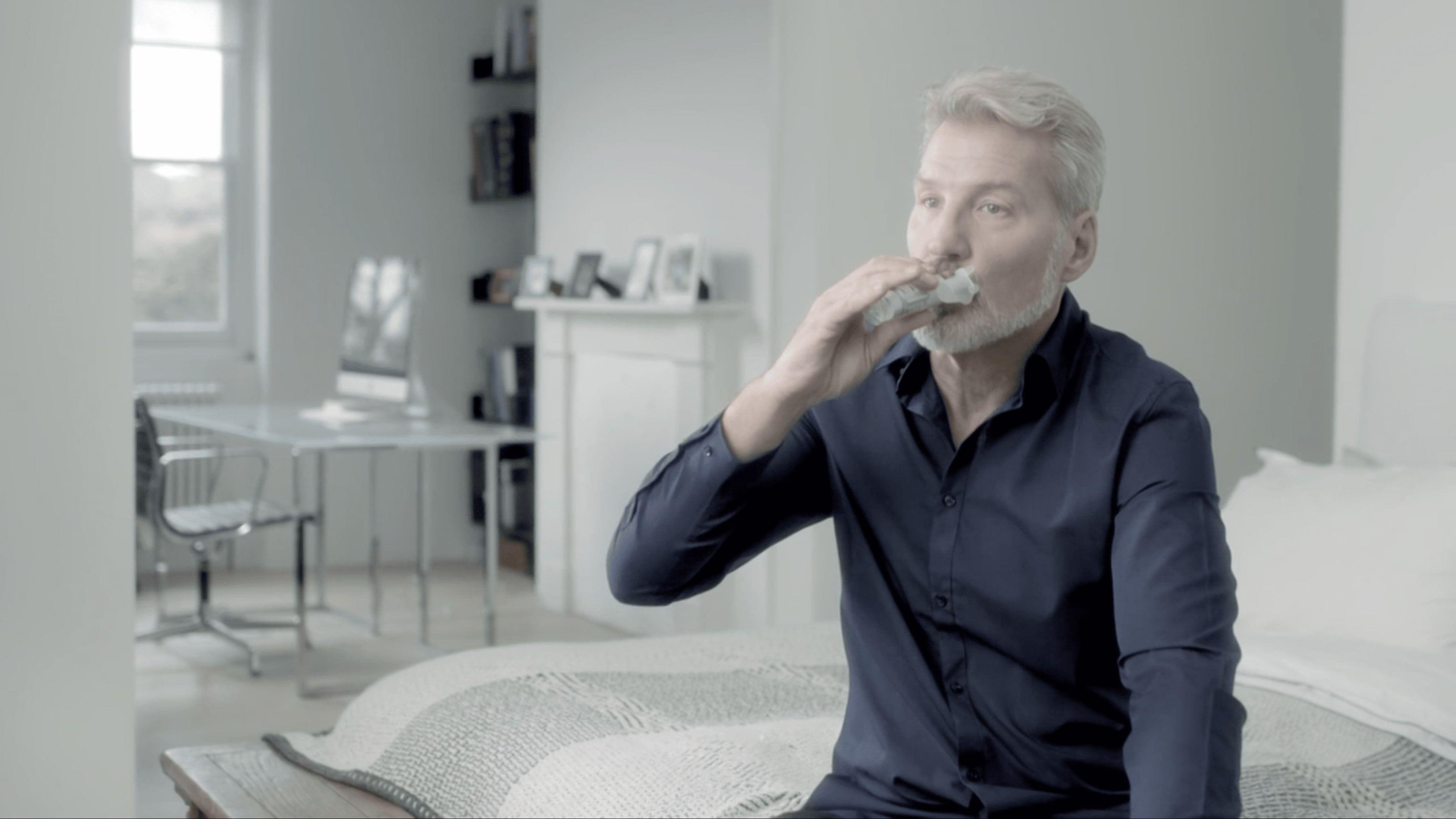 Muž sediaci na posteli drží v ruke inhalátor a vdychuje uvoľnenú dávku liečiba